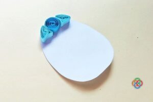 White egg template with blue quilled paper shapes arranged along the top edge, beginning the design of a quilled Easter egg.