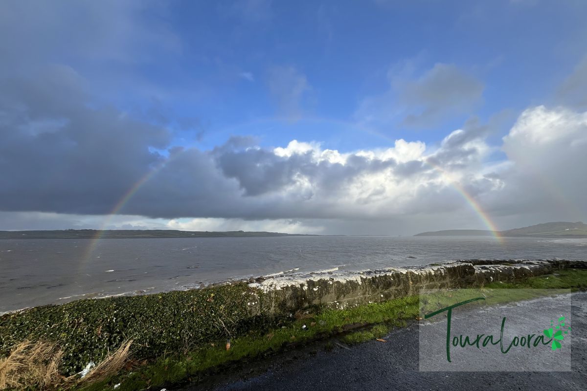 Rainbow stretching over a coastal landscape in the west of Ireland with ocean waves, stone wall, and cloudy sky.