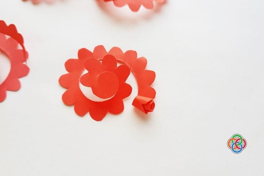 Red paper rosettes arranged in a spiral on a white surface, with a small colorful emblem in the bottom-right corner, suggesting crafting decorations for parties or events.