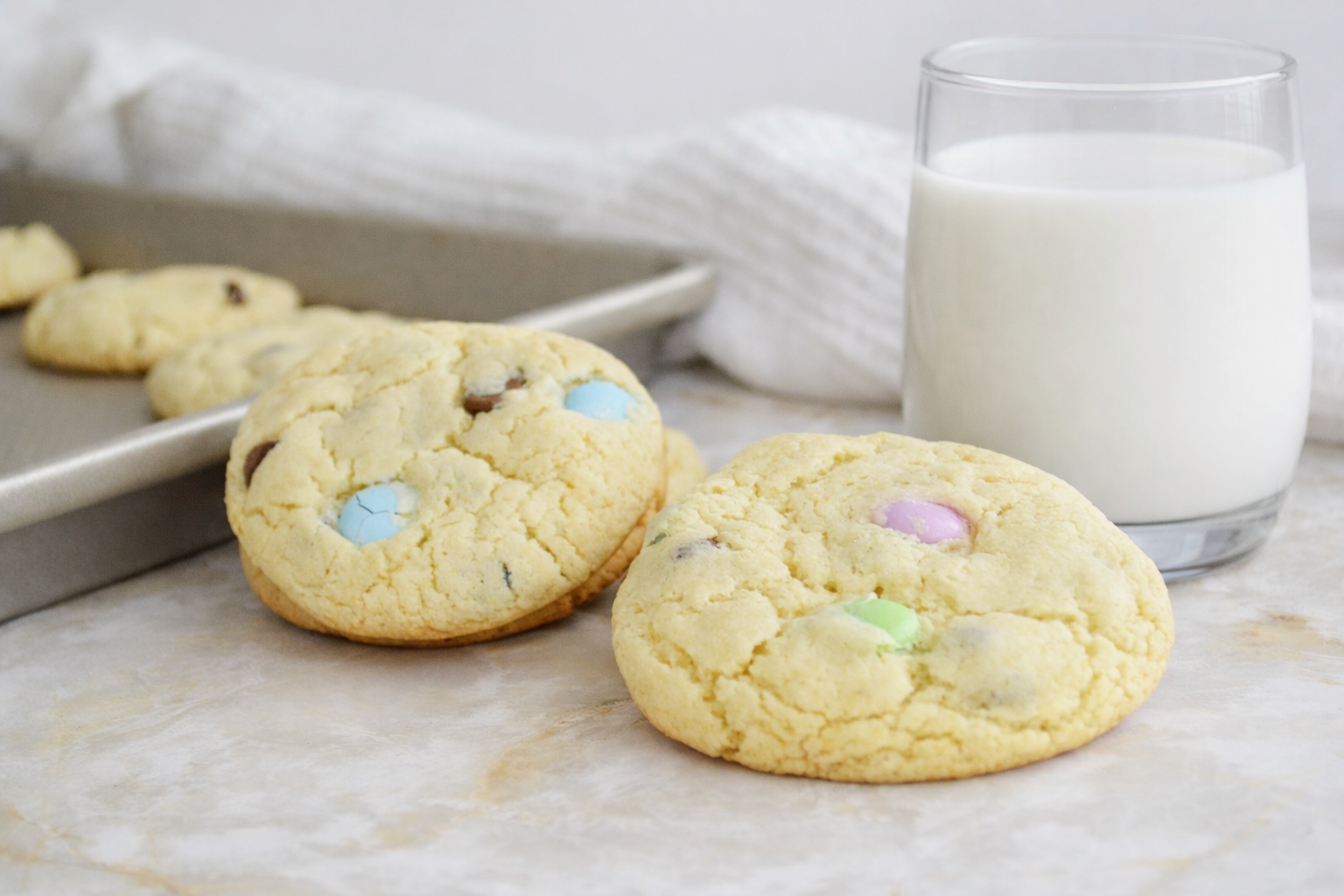 Two soft spring cake mix cookies with pastel M&M candies and chocolate chips on a marble surface, served with a glass of milk in the background.