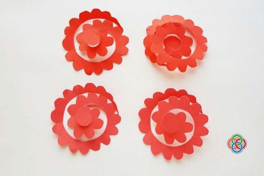 Four red spiral paper templates with scalloped edges arranged on a white background, ready to be rolled into paper roses for a wreath craft.