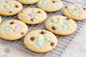 Freshly baked spring cake mix cookies with pastel M&M candies and chocolate chips cooling on a wire rack on a marble countertop.