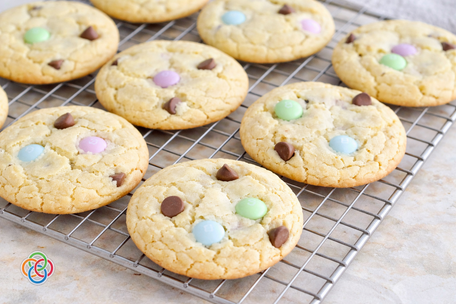 Freshly baked spring cake mix cookies with pastel M&M candies and chocolate chips cooling on a wire rack on a marble countertop.