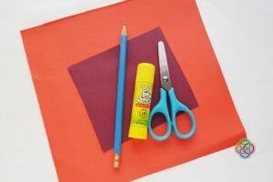 Craft supplies laid out: orange and purple paper, blue pencil, yellow glue stick, turquoise scissors on a white surface.