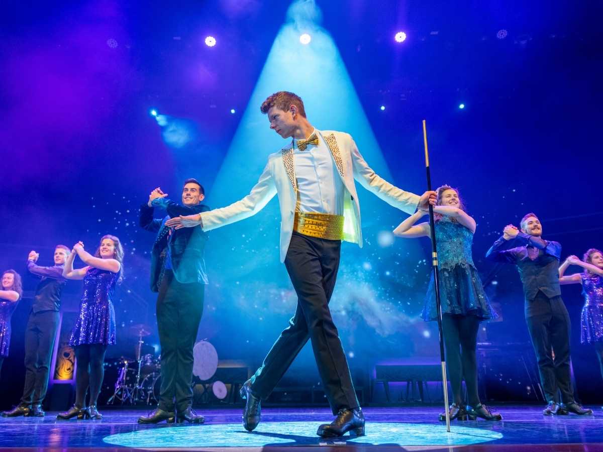 Celtic Throne Irish Dance Show | Irish American Mom Male dancer in a white jacket with gold belt and bow tie leads a stage performance, holding a baton as backup dancers in glittery outfits pose around him under blue spotlights.