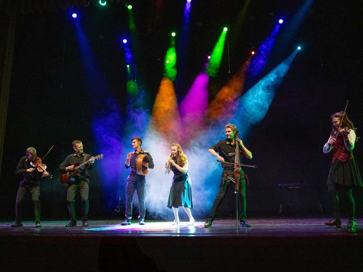 Celtic Throne Irish Dance and Entertainment | Irish American Mom Group of musicians and performers on stage during a Celtic-themed live show with colorful spotlights, including violinists, a guitarist, a cellist, and a singer performing Irish music.