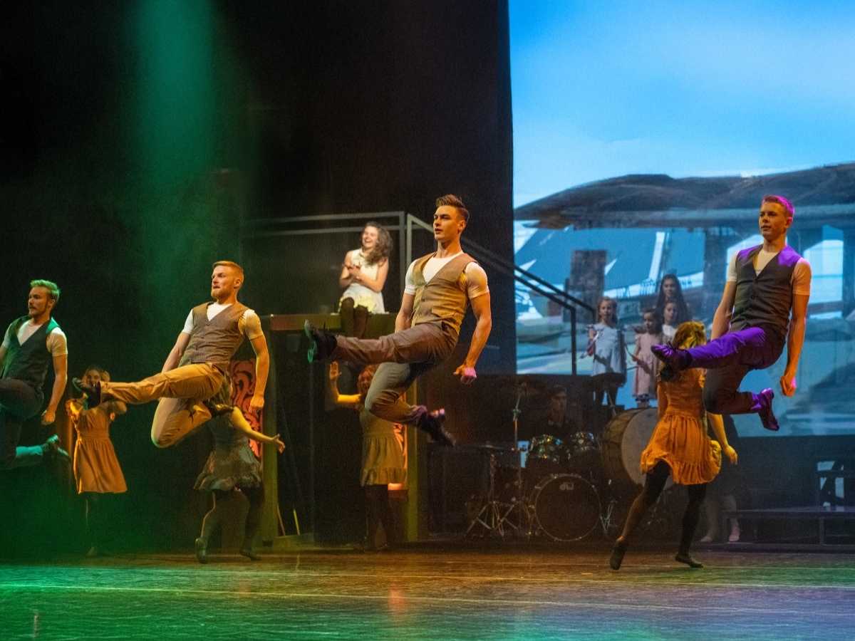High Stepping Celtic Throne Irish Danceing Theater Show USA Tour | Irish American Mom Dancers in tan outfits perform mid-air kicks on a stage, with a live band visible in the background under colorful lighting.