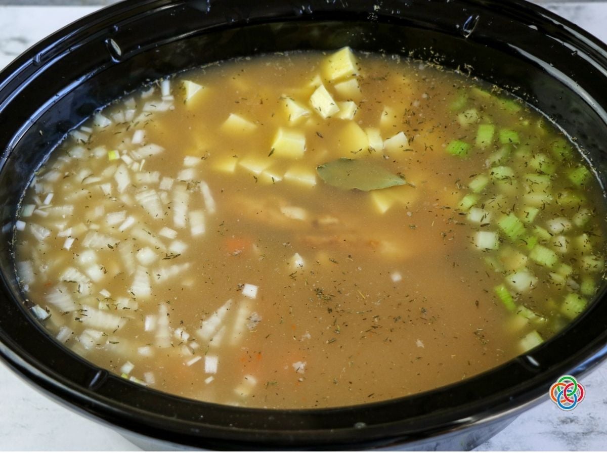 Adding broth and seasonings to Ingredients in a slow cooker for ham bone soup | Irish American Mom Slow cooker filled with broth, ham bone, potatoes, carrots, celery, onions, white beans, bay leaves and herbs ready to cook into ham bone soup