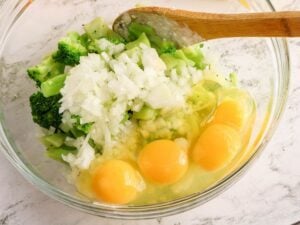 Adding onions to wet ingredients and broccoli for cheesy cornbread | Irish American Mom Chopped broccoli, onion and eggs being combined in a large glass mixing bowl while preparing cheesy broccoli cornbread, with shredded cheddar cheese and melted butter nearby.