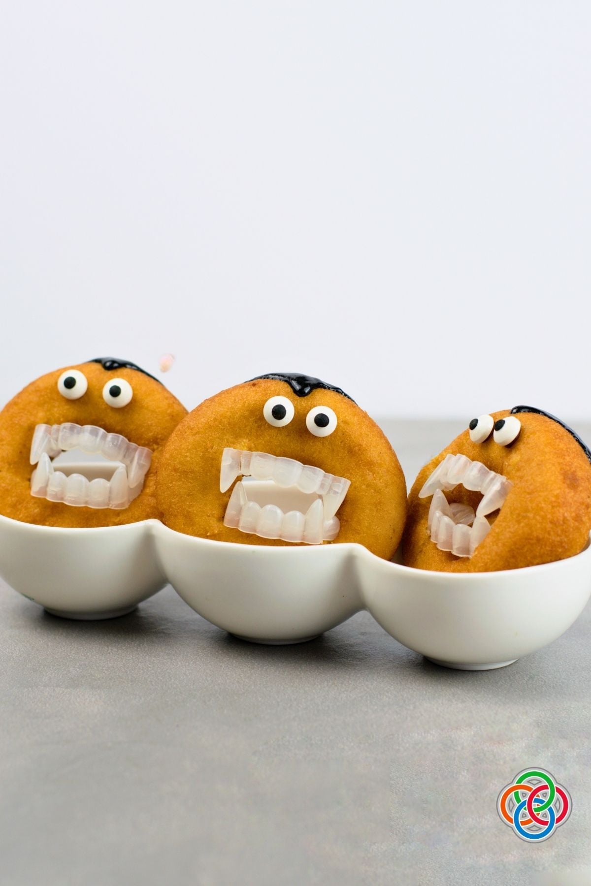 Three golden Dracula donuts decorated with candy eyes, black icing hairlines and plastic vampire fangs, displayed upright in white bowls.