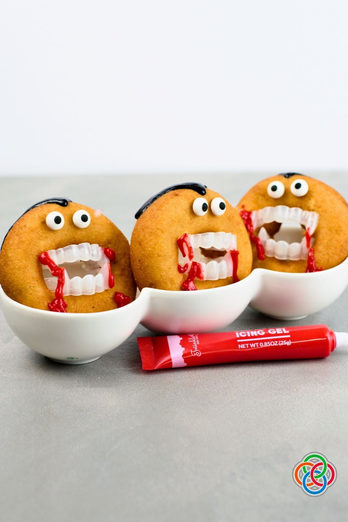 Three Dracula donuts decorated with candy eyes, black icing hair, plastic vampire fangs and dripping red icing to resemble blood, with a tube of red icing in front.