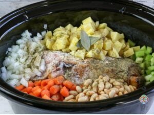 Adding seasonings to Ingredients in a slow cooker for ham bone soup | Irish American Mom Ham bone, diced potatoes, carrots, celery, onions and white beans in a slow cooker topped with bay leaves, thyme and seasonings before adding broth.