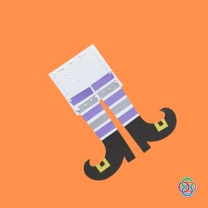 Applying cardboard support to a witch legs Halloween Bookmark | Irish American Mom Paper witch legs bookmark being assembled with glue and a white backing piece, featuring purple and gray striped stockings and curled black witch shoes with yellow buckles.