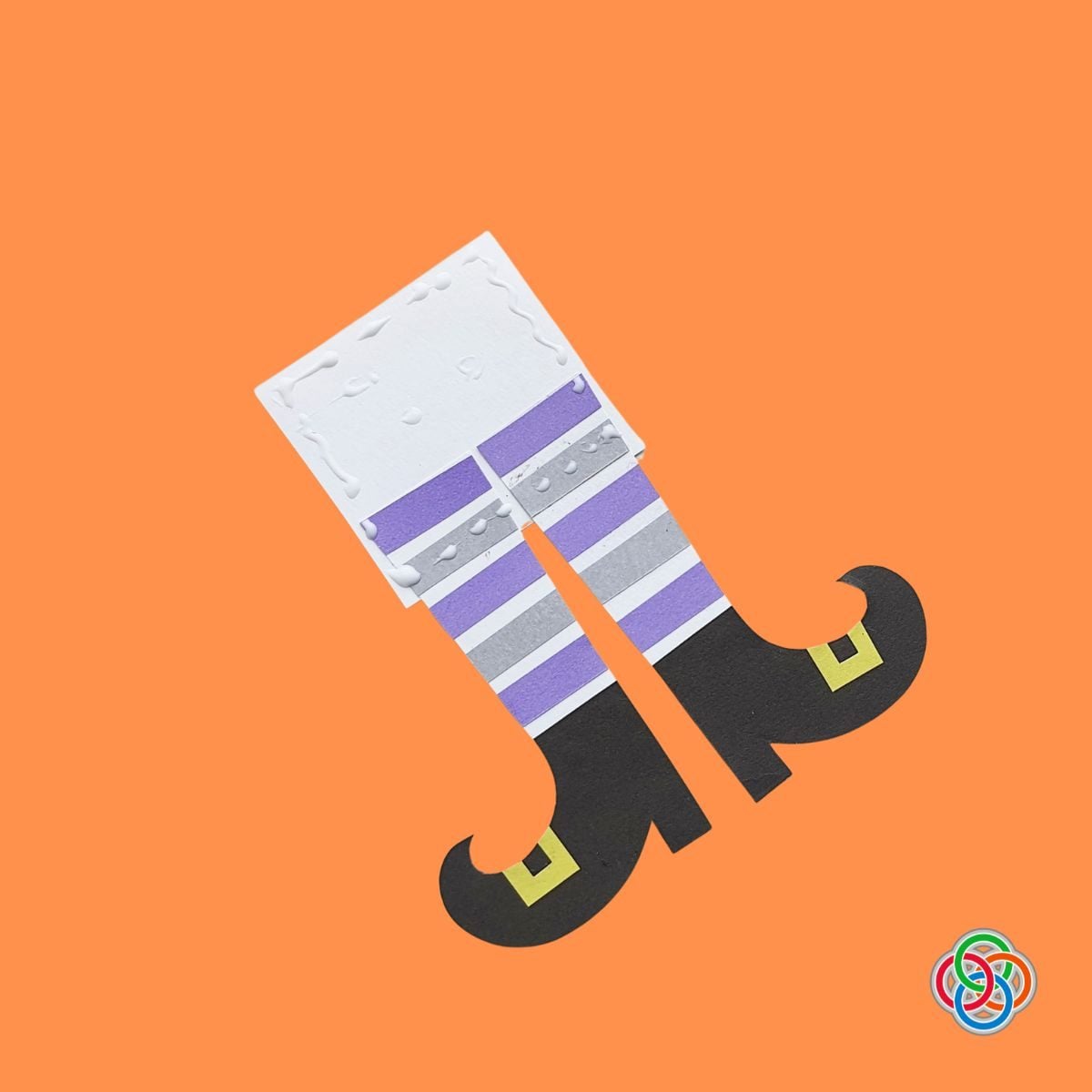 Applying cardboard support to a witch legs Halloween Bookmark | Irish American Mom Paper witch legs bookmark being assembled with glue and a white backing piece, featuring purple and gray striped stockings and curled black witch shoes with yellow buckles.