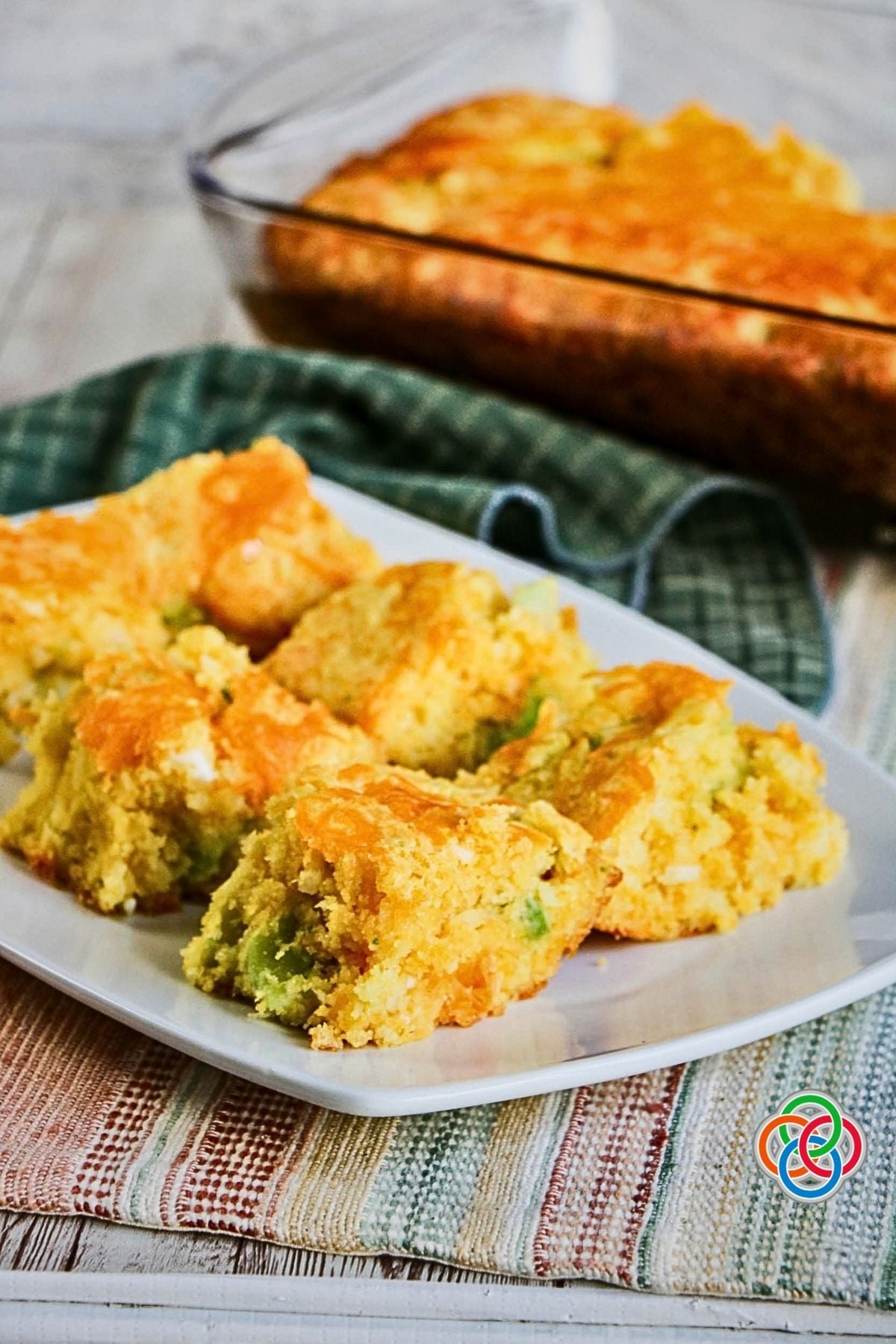 Broccoli Cheddar Cornbread Baked in a Casserole and Served in pieces on a platter | Irish American Mom Golden cheesy broccoli cornbread cut into squares and served on a white platter, with the remaining broccoli cheddar cornbread in a casserole dish in the background.