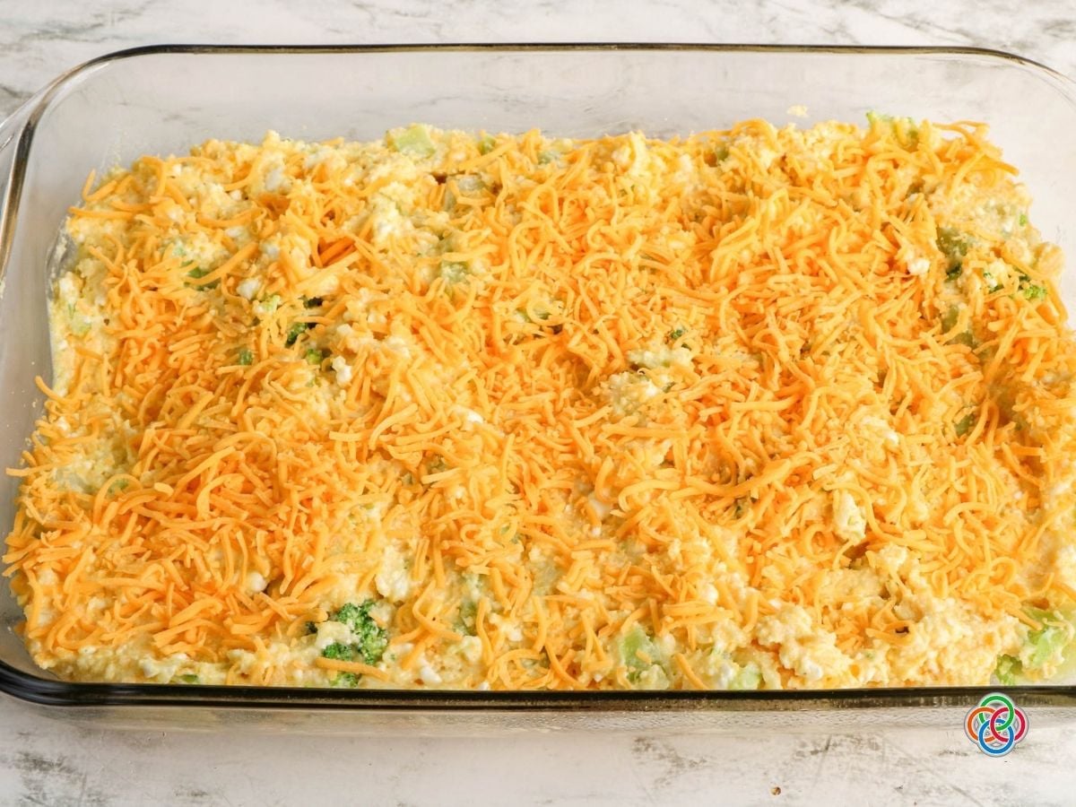 Cheddar broccoli cornbread batter in a casserole dish topped with grated cheddar ch | Irish American Mom Unbaked broccoli cornbread batter in a glass casserole dish topped generously with shredded cheddar cheese and ready for the oven.