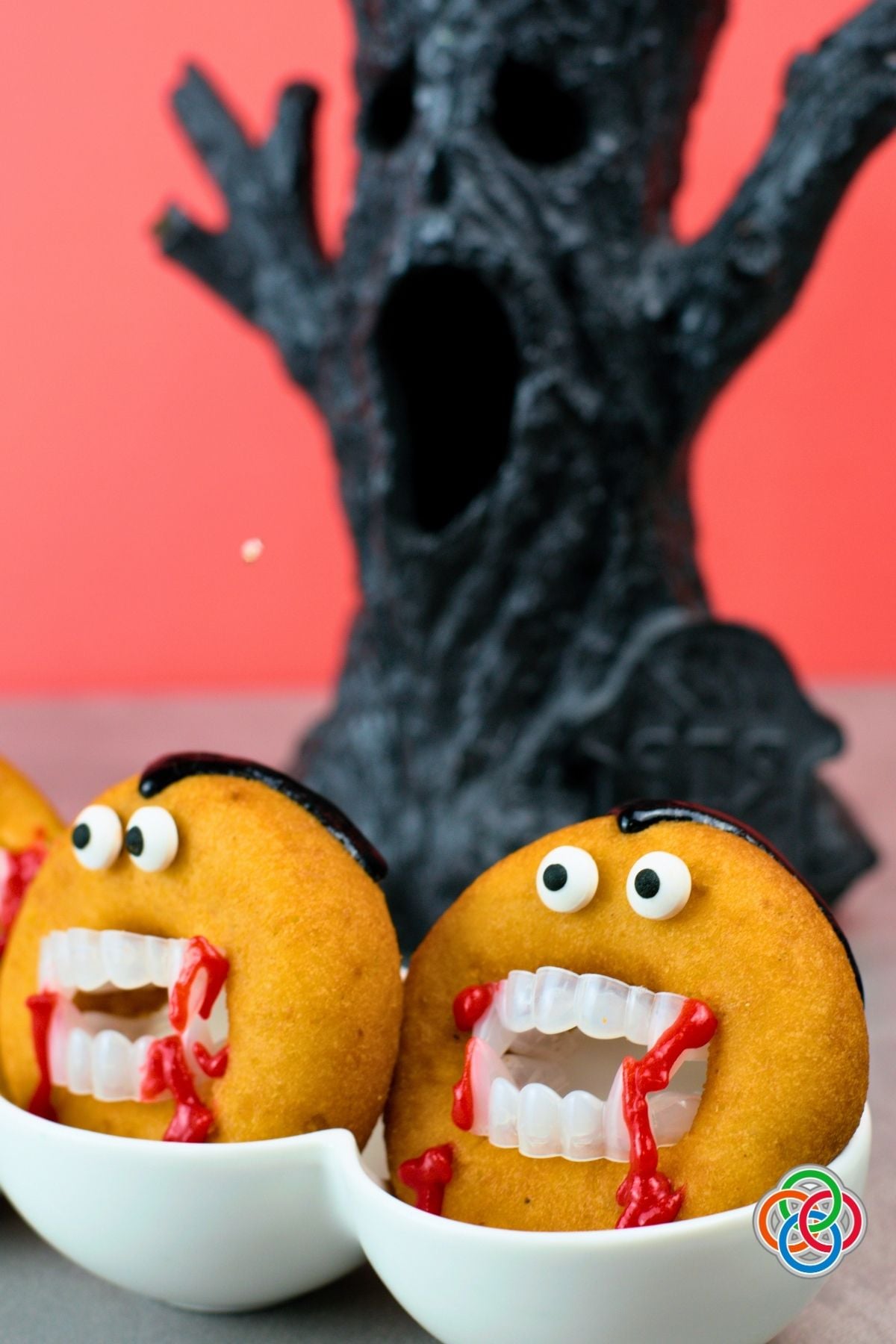 Two Dracula donuts with candy eyes, vampire teeth, black icing hair and dripping red icing displayed in white bowls in front of a spooky black Halloween tree.
