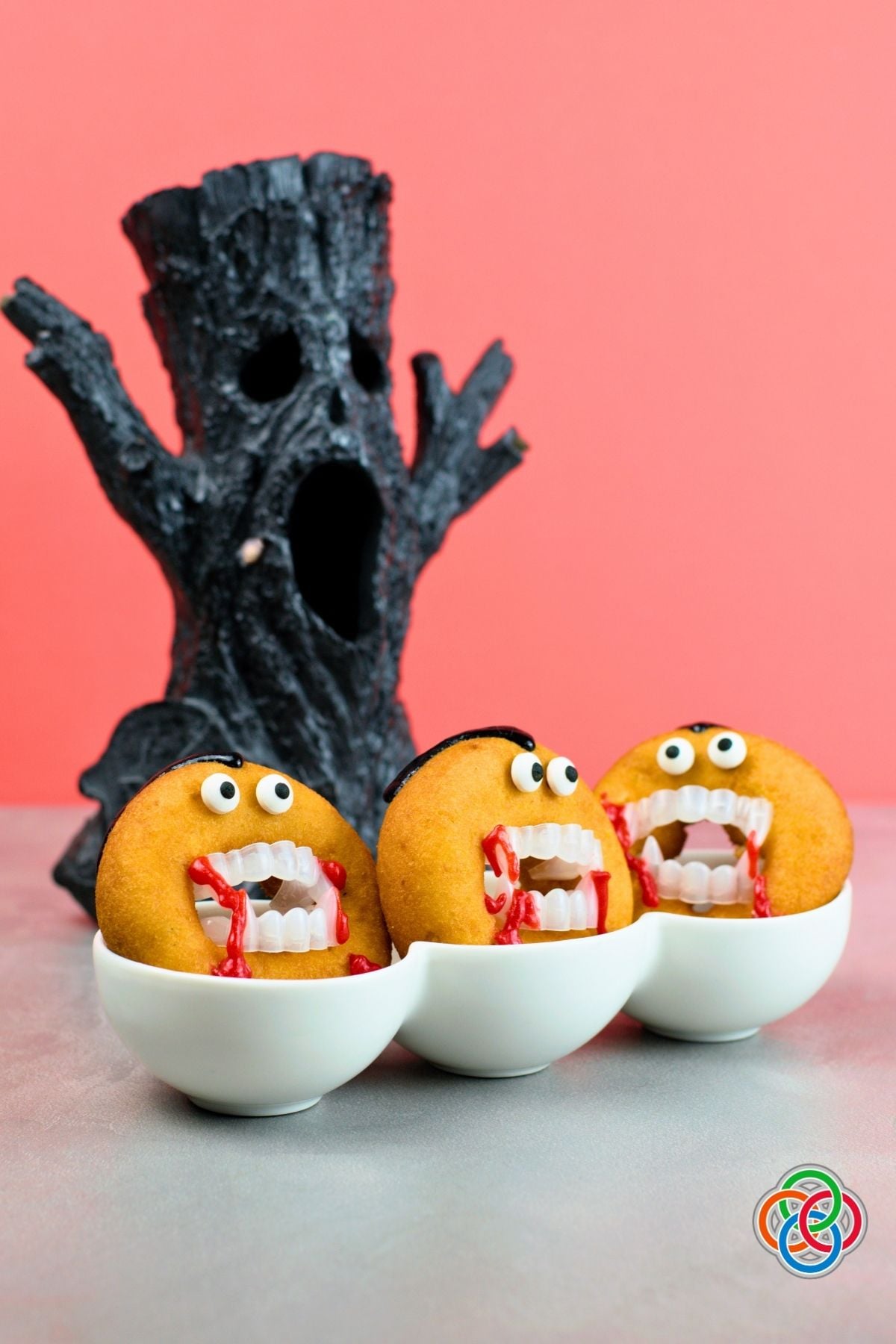 Three Dracula donuts decorated with candy eyes, vampire teeth, black icing hair and dripping red icing, displayed in white bowls beside a spooky black tree against an orange-red background.