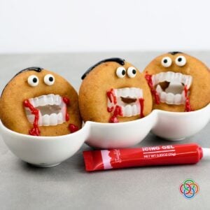 Three Dracula donuts decorated with candy eyes, black icing hair, plastic vampire fangs and dripping red icing to resemble blood, with a tube of red icing in front.