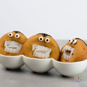 Three golden Dracula donuts decorated with candy eyes, black icing hairlines and plastic vampire fangs, displayed upright in white bowls.
