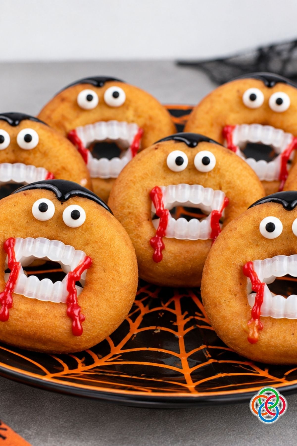 Dracula donuts decorated with candy eyes, black icing hair, vampire teeth and dripping red icing arranged on a black and orange spiderweb Halloween plate.