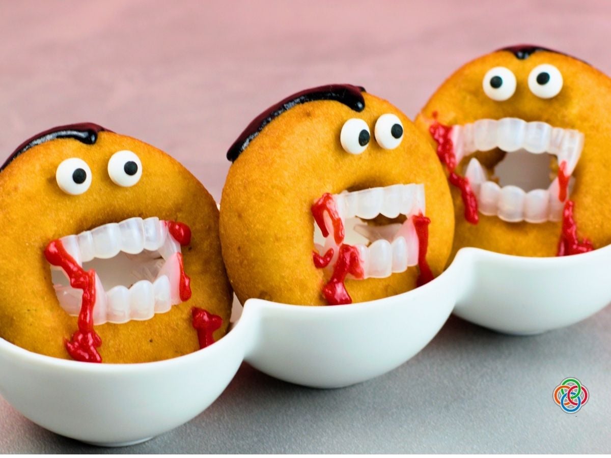 Close-up of three Dracula donuts decorated with candy eyes, black icing hair, plastic vampire fangs and dripping red icing, displayed upright in white bowls.