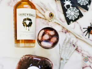 Overhead view of a dark grape juice and peanut butter whiskey Halloween cocktail with ice, surrounded by a whiskey bottle, skeleton hands, eyeball decoration and Halloween spider.