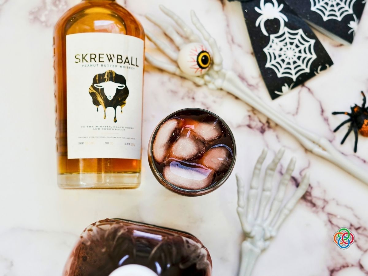 Overhead view of a dark grape juice and peanut butter whiskey Halloween cocktail with ice, surrounded by a whiskey bottle, skeleton hands, eyeball decoration and Halloween spider.
