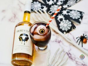 Peanut butter whiskey and grape juice cocktail served over ice with a decorative Halloween eyeball and striped straw, beside a whiskey bottle and white skeleton hand.