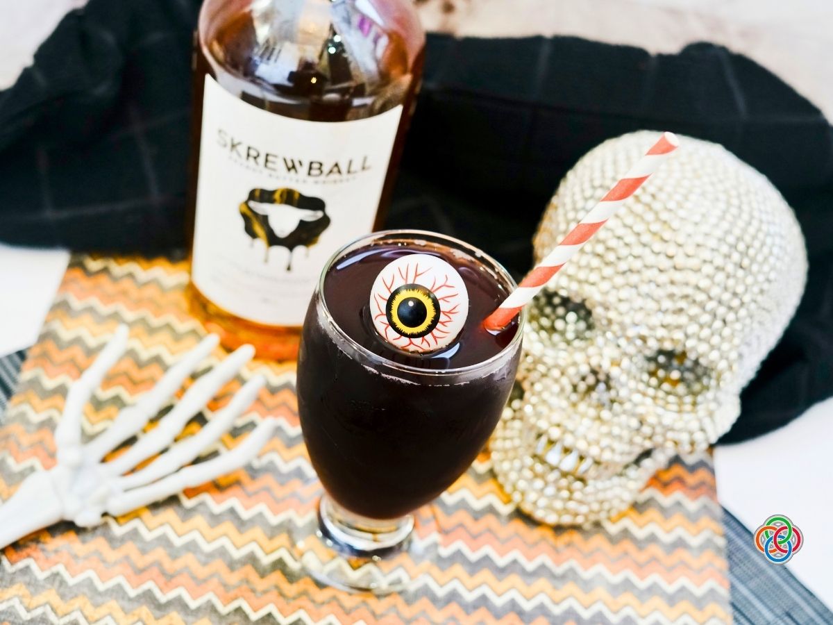 Dark purple Halloween cocktail topped with a decorative eyeball and striped straw beside a peanut butter whiskey bottle, jeweled skull and skeleton hand.