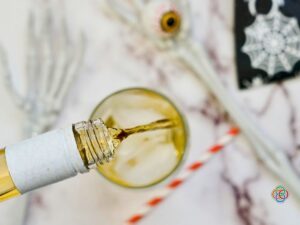 Peanut butter whiskey being poured from a bottle into a glass with ice while Halloween skeleton hands, a decorative eyeball and spider web napkins decorate the background.
