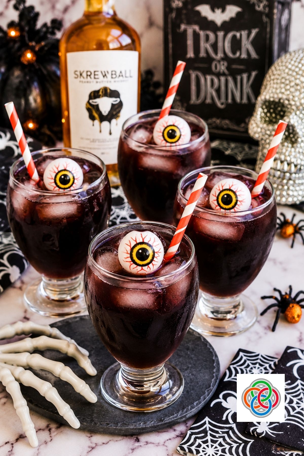 Four dark purple Halloween cocktails made with peanut butter whiskey and grape juice, served over ice with creepy eyeball garnishes and red striped straws, surrounded by a jeweled skull, skeleton hand, spiders and Halloween decorations.
