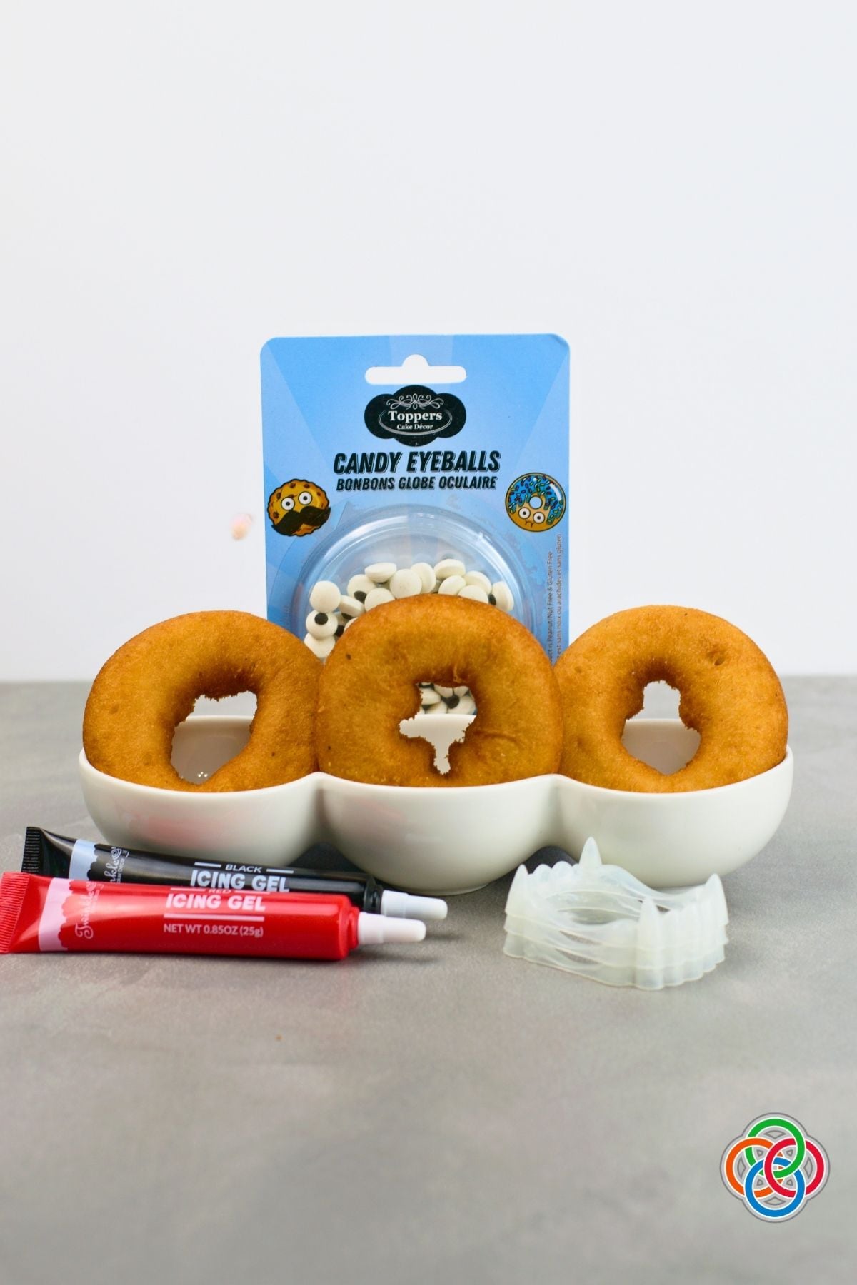 Ingredients and supplies for Dracula donuts including three plain donuts, candy eyeballs, plastic vampire teeth, and tubes of red and black icing gel.