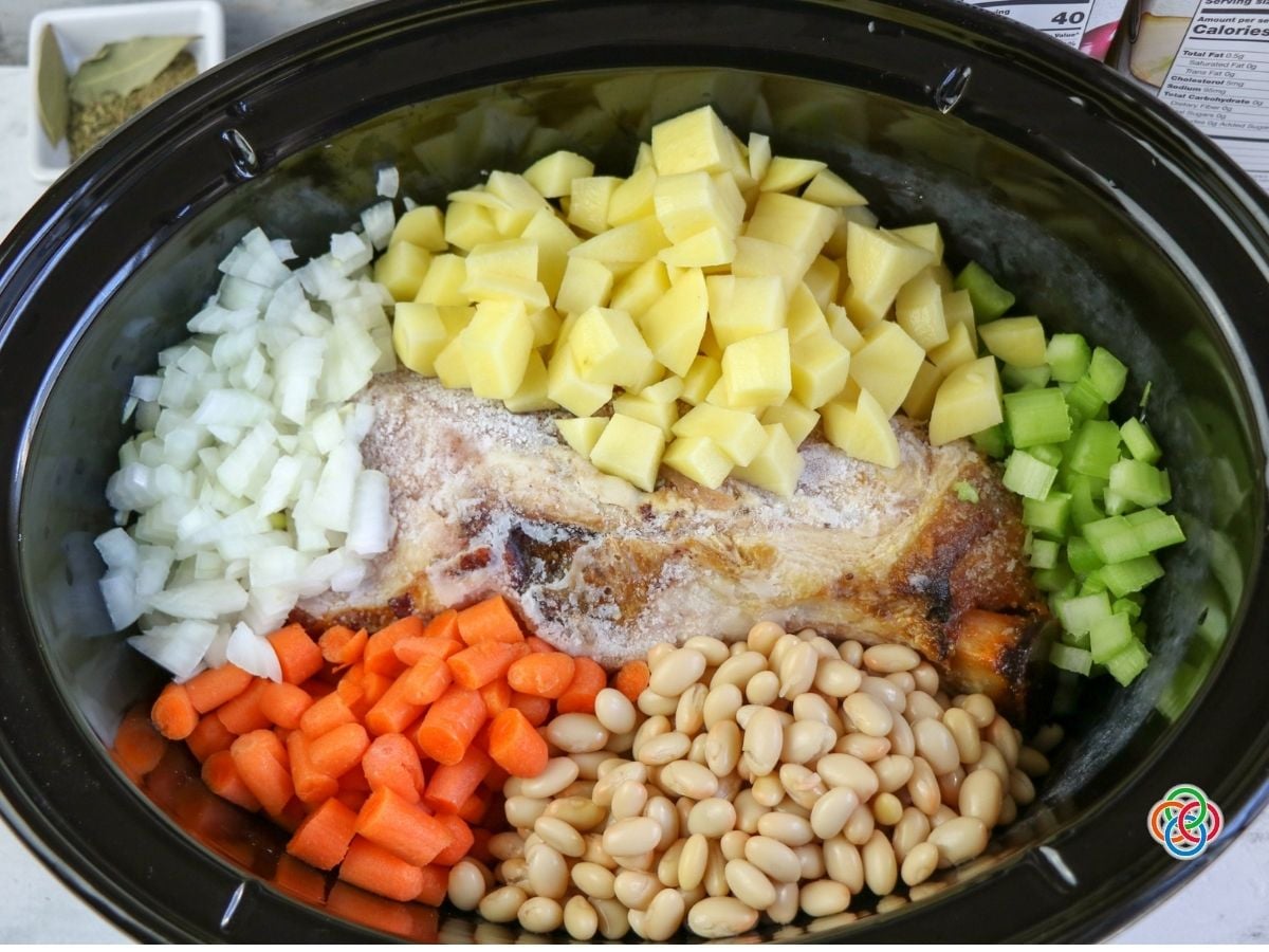 Ingredients in a slow cooker ham bone soup | Irish American Mom Ham bone in a slow cooker surrounded by diced potatoes, carrots, celery, onions and white beans, ready to make homemade ham bone soup.