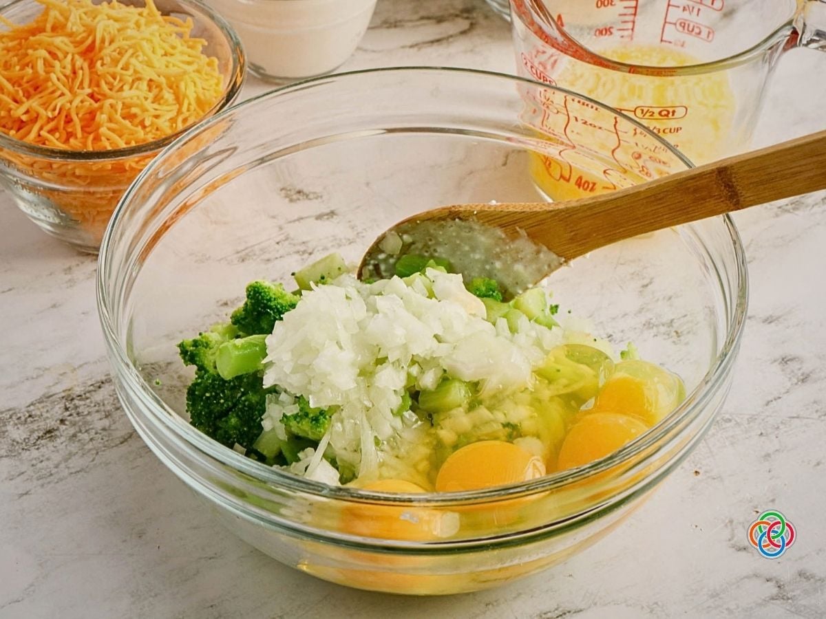 Mixing wet ingredients and broccoli for cheesy cornbread | Irish American Mom Chopped broccoli, onion and eggs being combined in a large glass mixing bowl while preparing cheesy broccoli cornbread, with shredded cheddar cheese and melted butter nearby.