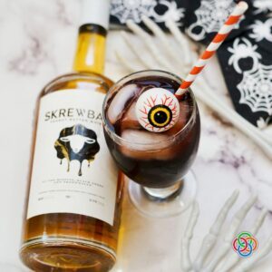Halloween eyeball cocktail made with peanut butter whiskey and grape juice, served over ice with a red striped straw and decorative eyeball, surrounded by spooky skeleton hands and Halloween napkins.
