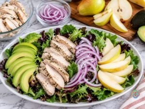 Pear Harvest Salad How to Assemble | Irish American Mom Harvest pear salad being assembled on a white platter with spring greens, sliced Bartlett pears, avocado, cooked turkey breast and thinly sliced red onion.