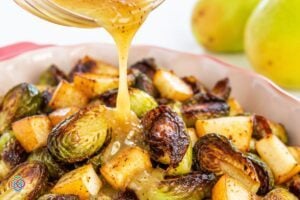 Pouring a maple syrup and mustard glaze on roasted Brussels sprouts | Irish American Mom Maple Dijon dressing being poured over crispy roasted Brussels sprouts and golden pear pieces in a red serving dish, with fresh pears in the background.