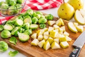 Preparing Brussels sprouts and pears for roasting | Irish American Mom Halved Brussels sprouts and chopped skin-on pears on a wooden cutting board with whole pears, a kitchen knife and a red and white checkered cloth.