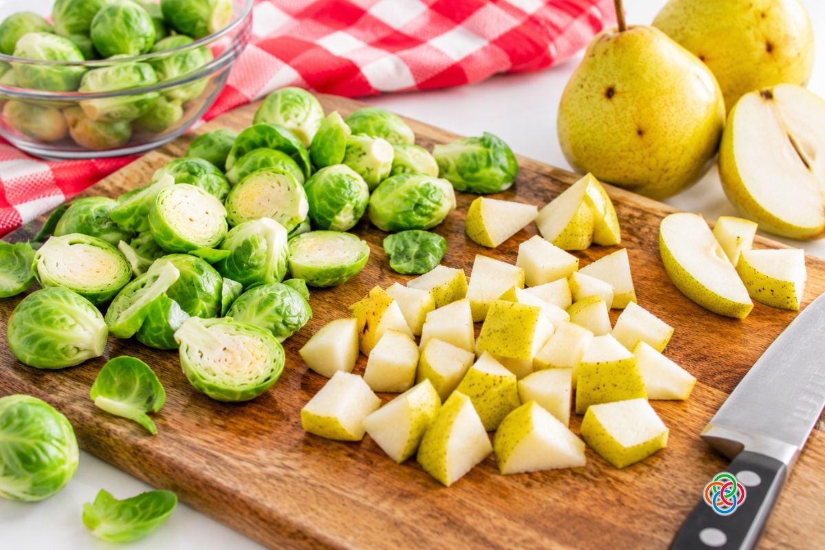 Preparing Brussels sprouts and pears for roasting | Irish American Mom Halved Brussels sprouts and chopped skin-on pears on a wooden cutting board with whole pears, a kitchen knife and a red and white checkered cloth.