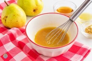 Preparing a maple syrup and mustard glaze for Brussels sprouts | Irish American Mom Maple syrup, Dijon mustard and apple cider vinegar being whisked together in a white bowl with a red rim, surrounded by fresh pears and a red and white checkered cloth.