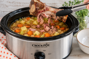 Removing the ham bone from the crock pot | Irish American Mom Removing a large meat bone from a slow cooker full of cooked soup with vegetables.