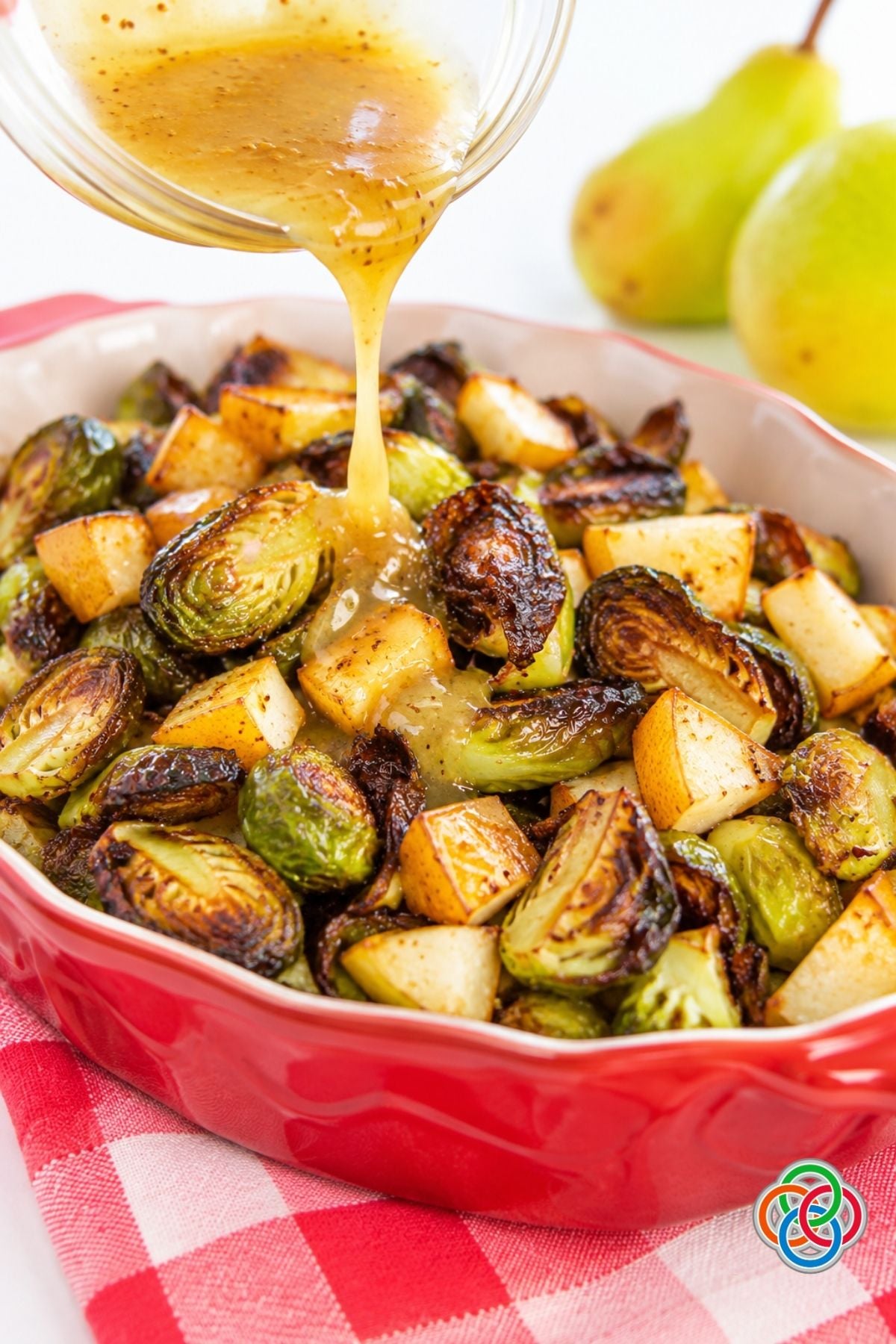 Roasted Brussels Sprouts and Pears with Maple Dijon Dressing | Irish American Mom Maple Dijon dressing being poured over crispy roasted Brussels sprouts and golden pear pieces in a red serving dish, with fresh pears in the background.