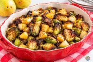 Roasted Brussels sprouts and pears with maple mustard glaze in a large serving dish | Irish American Mom Roasted Brussels sprouts and golden pear pieces with caramelized crispy edges and maple Dijon glaze served in a large red ceramic serving dish on a red and white checkered cloth.