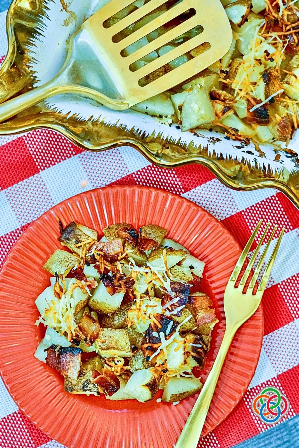 Serving of Parmesan Bacon Roast Potatoes | Irish American Mom Bacon Parmesan roasted potatoes with crispy bacon, garlic and golden Parmesan served on a red plate beside a gold fork, with more roasted potatoes in the background.