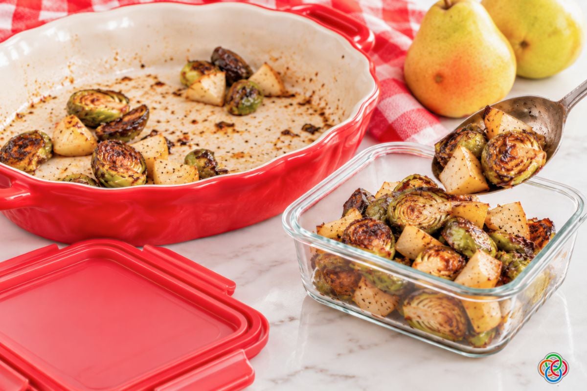 Storing leftover Brussels sprouts and pears | Irish American Mom Leftover roasted Brussels sprouts and pears being spooned from a nearly empty red serving dish into a rectangular glass storage container with a matching red lid beside it.