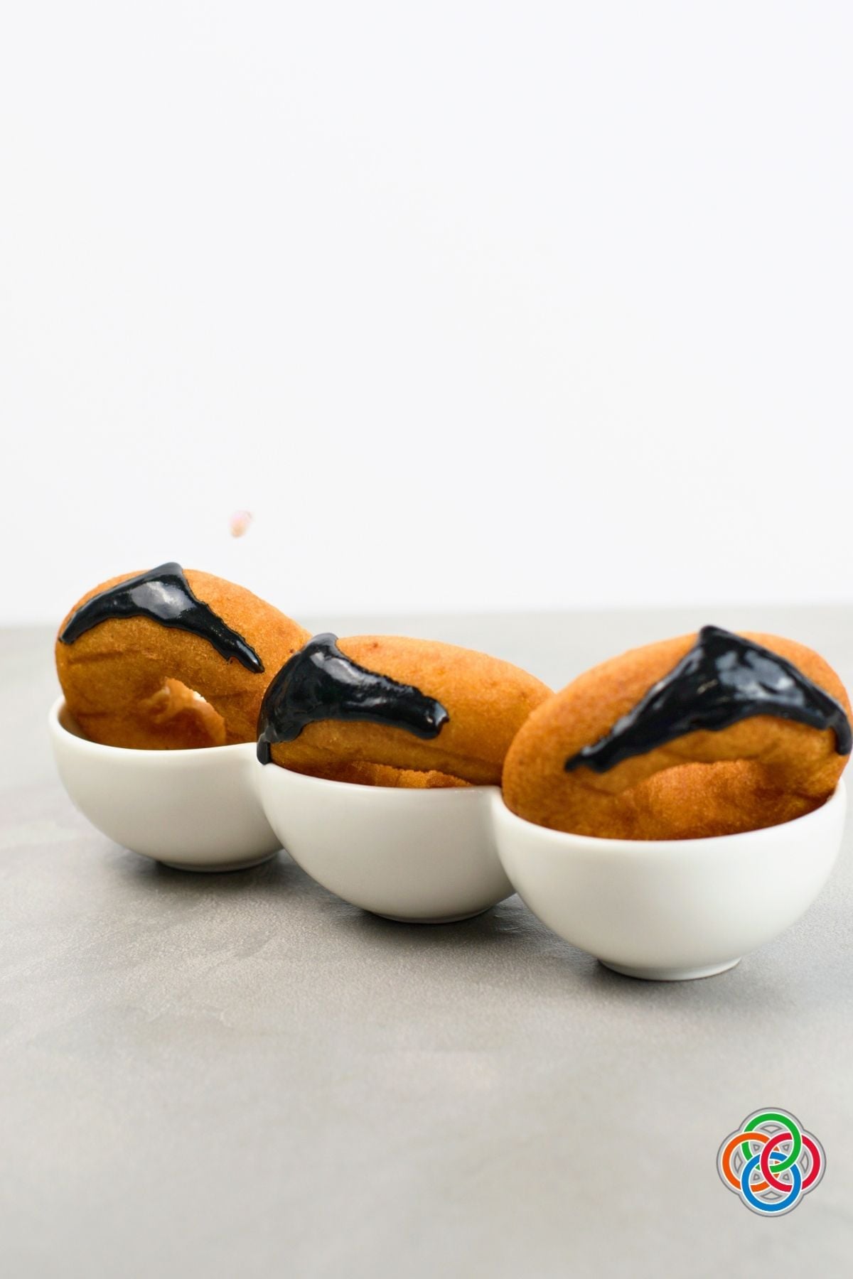 Three plain donuts in white bowls decorated with glossy black icing in a V-shaped widow’s peak to create Dracula’s hairline for a Halloween donut craft.