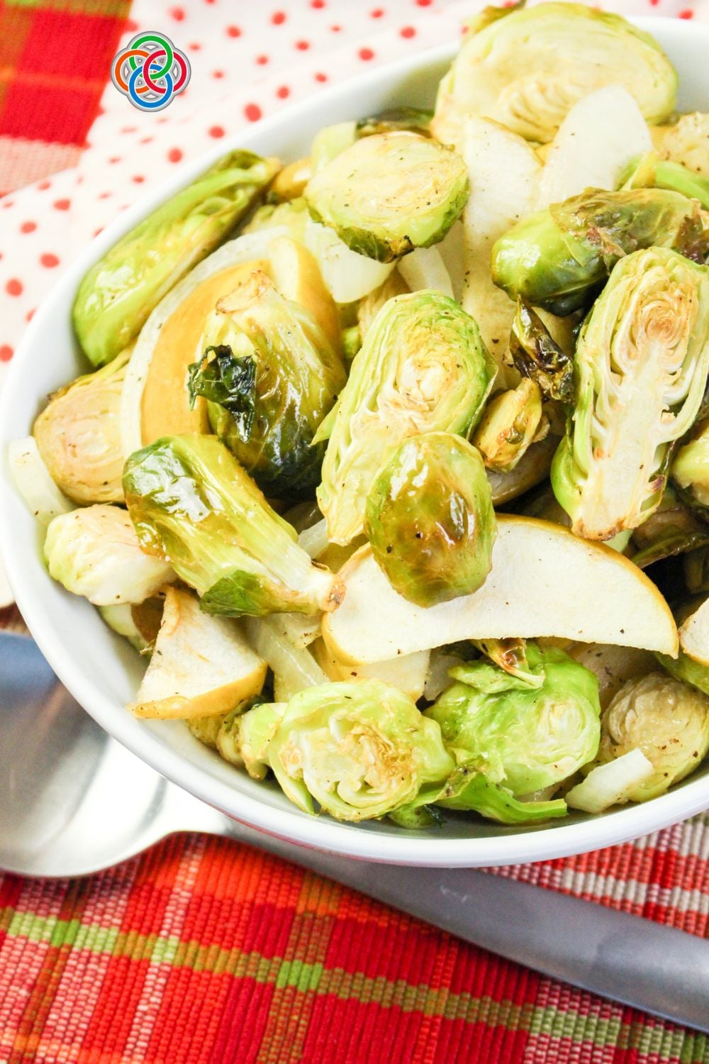 Close-up of roasted Brussels sprouts with sliced Granny Smith apples and sweet onions in a white serving bowl, arranged on a red plaid tablecloth with a red polka-dot napkin.