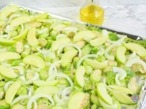 Fresh halved Brussels sprouts, sliced green Granny Smith apples, and sweet onion rings spread across a foil-lined baking sheet, ready for roasting. A glass bottle of olive oil sits behind the tray on a white marble countertop.