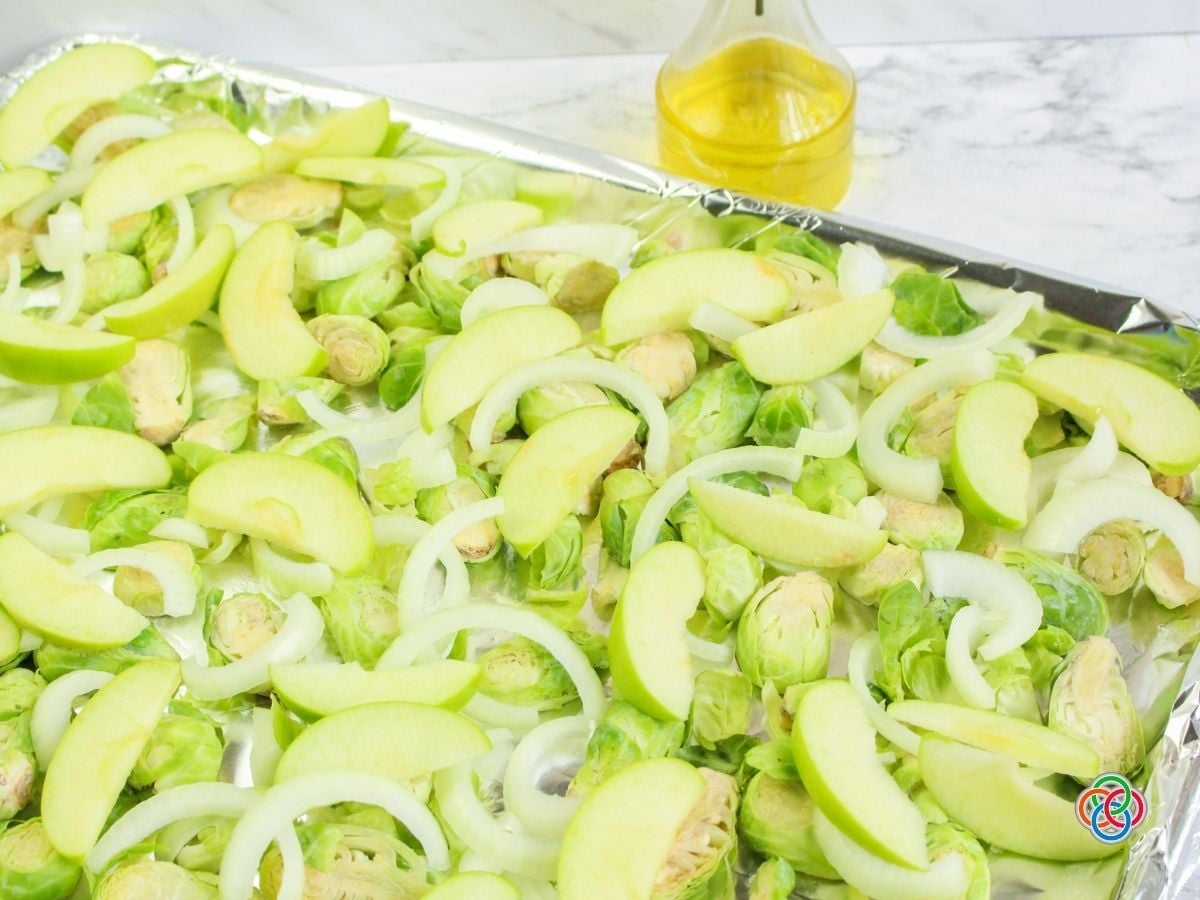 Fresh halved Brussels sprouts, sliced green Granny Smith apples, and sweet onion rings spread across a foil-lined baking sheet, ready for roasting. A glass bottle of olive oil sits behind the tray on a white marble countertop.